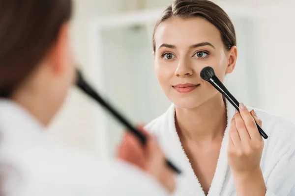 Улыбающаяся молодая женщина смотрит в зеркало и наносит макияж в ванной комнате — стоковое фото