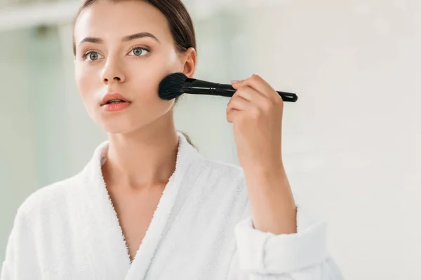 Beautiful girl in bathrobe applying bronzer with brush — Stock Photo