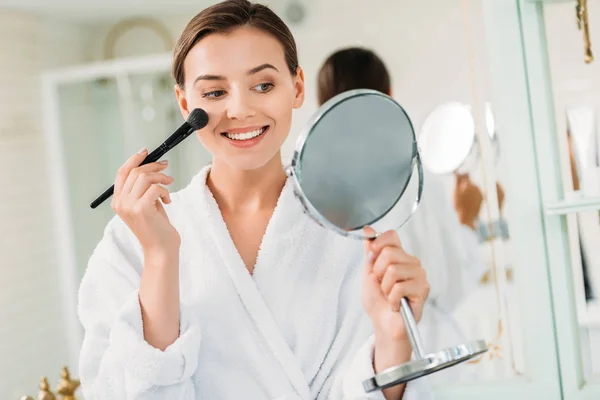 Heureuse jeune femme en peignoir tenant miroir et appliquant le maquillage dans la salle de bain — Photo de stock