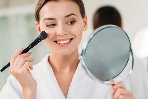 Hermosa mujer joven sonriente sosteniendo el espejo y aplicando maquillaje - foto de stock