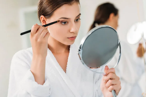 Hermosa joven mujer sosteniendo espejo y aplicando sombra de ojos - foto de stock