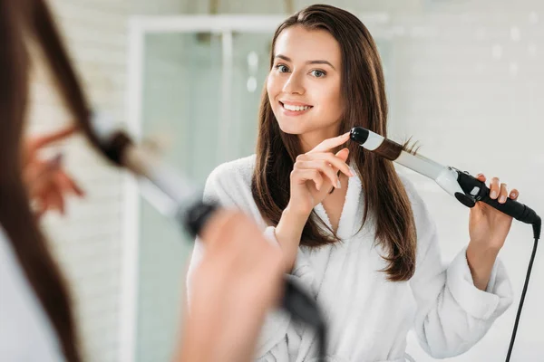 Menina morena feliz em roupão de banho usando frisador de cabelo no espelho no banheiro — Fotografia de Stock