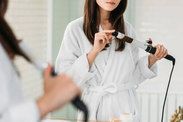 Tiro recortado de mujer joven en albornoz usando rizador de pelo en el espejo en el baño - foto de stock