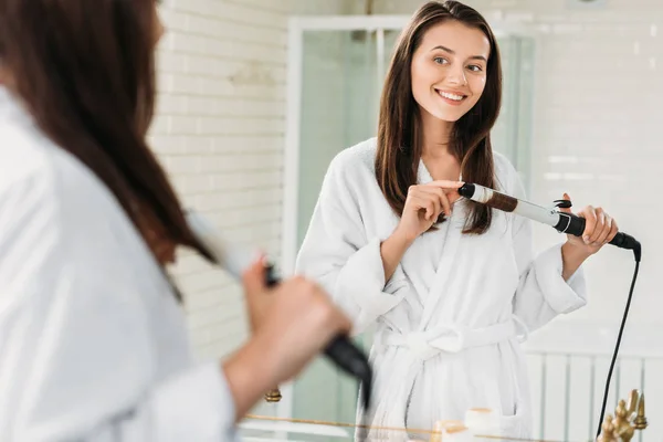 Hermosa mujer joven sonriente en albornoz usando rizador de pelo en el espejo en el baño - foto de stock