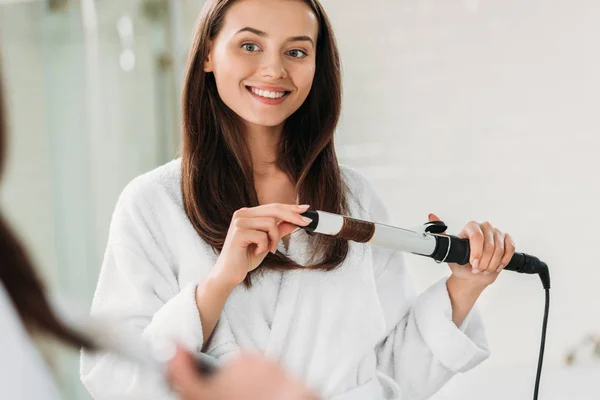 Sorrindo jovem mulher em roupão de banho usando frisador de cabelo no banheiro — Fotografia de Stock