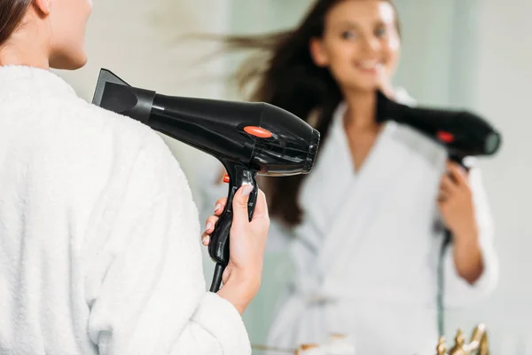 Selective focus of smiling girl in bathrobe using hair dryer at mirror in bathroom — Stock Photo