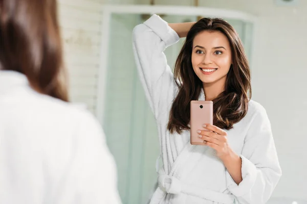 Belle jeune femme souriante en peignoir prenant selfie avec smartphone dans la salle de bain — Photo de stock