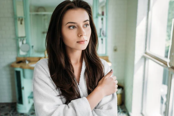 Стомлена брюнетка дівчина в халаті дивиться на вікно у ванній — стокове фото
