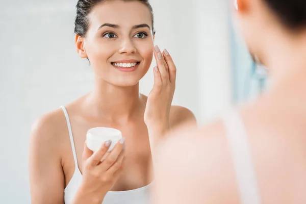 Hermosa sonriente joven aplicación de crema facial y mirando el espejo - foto de stock