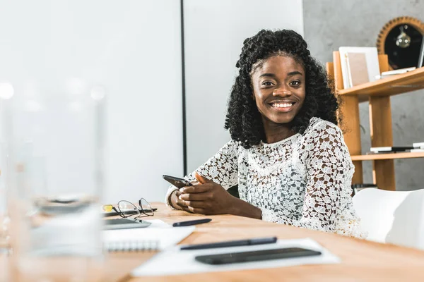 Retrato de mulher de negócios afro-americana sorridente com smartphone em mãos olhando para a câmera enquanto sentado no local de trabalho — Fotografia de Stock