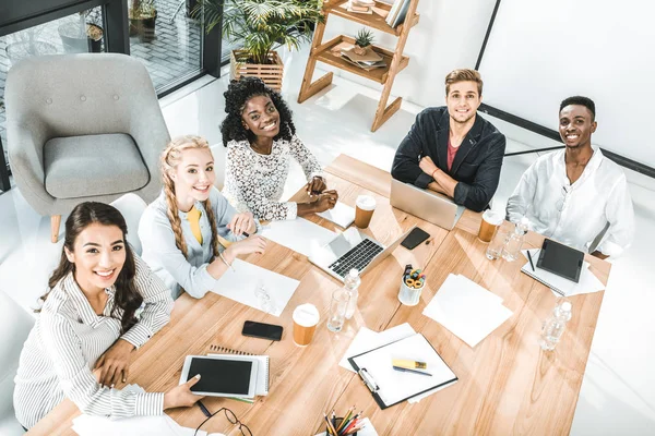 High angle view of multicultural group of business people looking at camera while sitting at table in conference room — Stock Photo