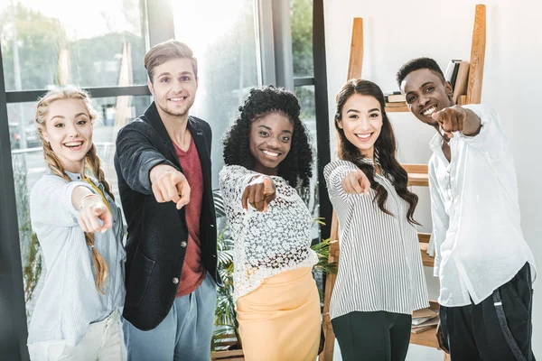 Multiethnic smiling business people pointing at camera while standing together in office — Stock Photo