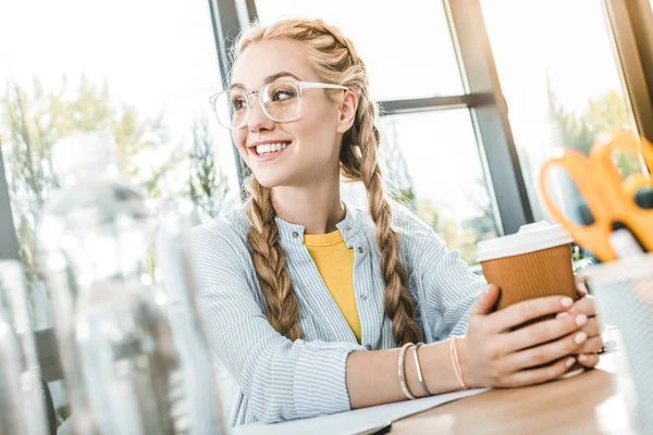Portrait de femme d'affaires souriante dans des lunettes avec du café pour aller dans les mains assis à table au bureau — Photo de stock
