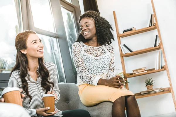 Multiethnic smiling businesswomen drinking coffee together during break in office — Stock Photo