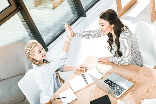 High angle view of multicultural businesswomen giving high five at workplace with laptop and notebooks — Stock Photo