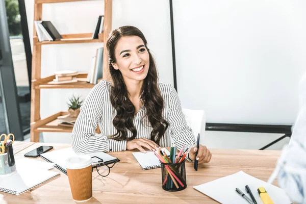Портрет улыбающейся азиатской предпринимательницы, сидящей на рабочем месте с различными офисными принадлежностями — стоковое фото