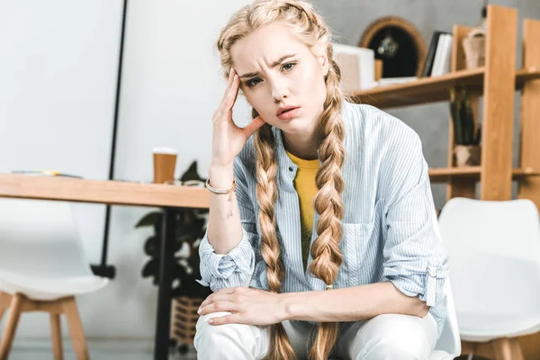 Portrait of young pensive businesswoman sitting at workplace in office and looking at camera — Stock Photo