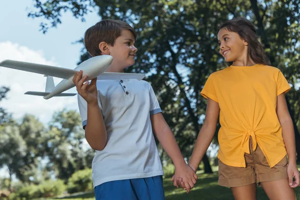 Низький кут зору щасливих дітей, що тримають модель літака та посміхаються один одному, тримаючись за руки та ходячи разом у парку — стокове фото