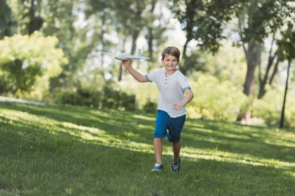 Adorável menino feliz segurando avião de brinquedo e sorrindo para a câmera no parque — Fotografia de Stock