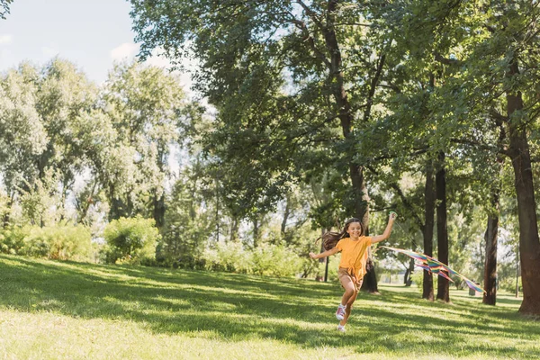 Lindo niño feliz jugando con colorido cometa y corriendo sobre hierba en parque - foto de stock