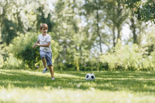 Милый мальчик бегает по траве и играет с футбольным мячом в парке — стоковое фото
