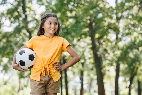 Lindo niño feliz sosteniendo pelota de fútbol y mirando hacia otro lado en el parque - foto de stock