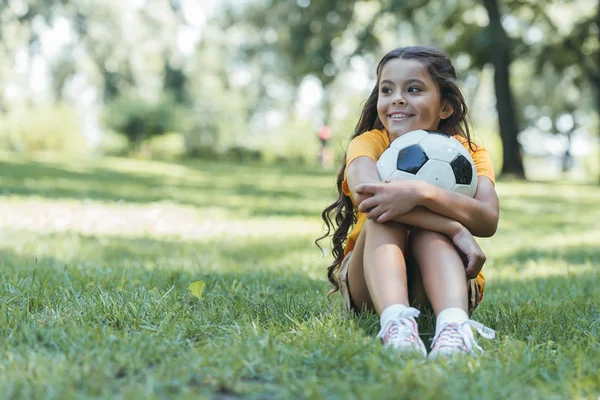 Adorable niño feliz sosteniendo pelota de fútbol y mirando hacia otro lado mientras está sentado en el parque - foto de stock