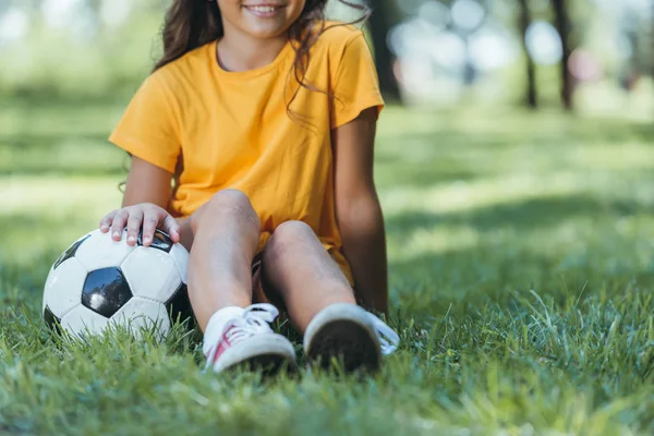 Recortado disparo de lindo niño sonriente sentado en la hierba con pelota de fútbol - foto de stock