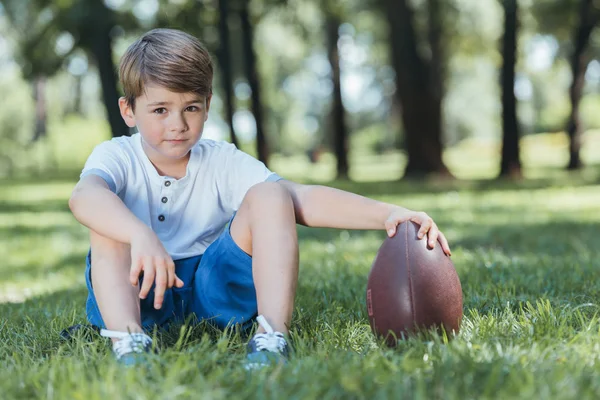 Lindo niño sosteniendo pelota de rugby mientras está sentado en la hierba y mirando a la cámara - foto de stock