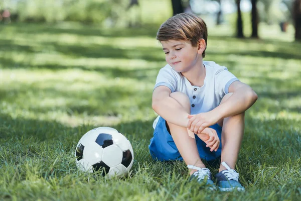 Милий маленький хлопчик сидить на траві і дивиться на футбольний м'яч — стокове фото