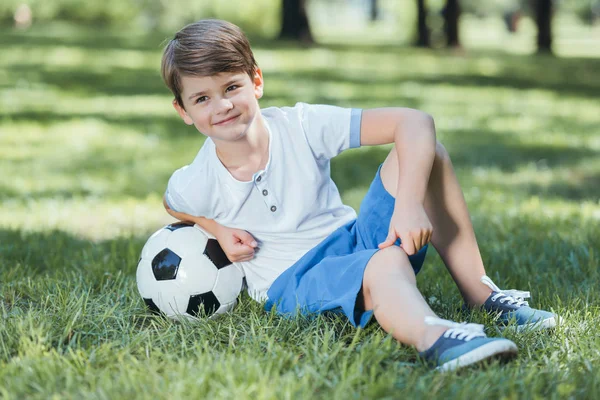 Adorable happy little boy resting in grass with soccer ball — Stock Photo