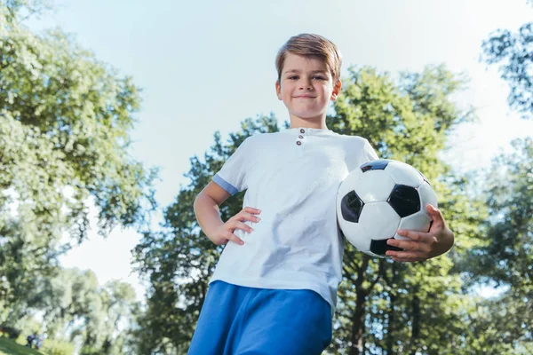 Visão de baixo ângulo do menino segurando bola de futebol e sorrindo para a câmera no parque — Fotografia de Stock