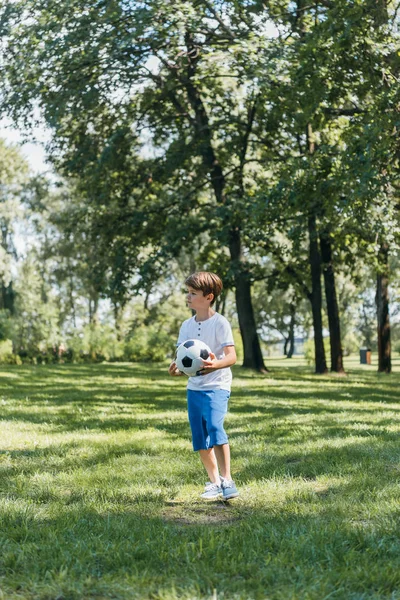 Lindo niño sosteniendo pelota de fútbol y mirando hacia otro lado en el parque - foto de stock