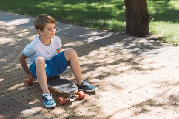 Vista ad alto angolo del ragazzo sorridente seduto sullo skateboard e guardando lontano nel parco — Foto stock