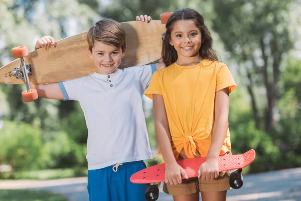 Симпатичные счастливые дети, держащие в руках скейтборды и улыбающиеся камерам в парке — стоковое фото