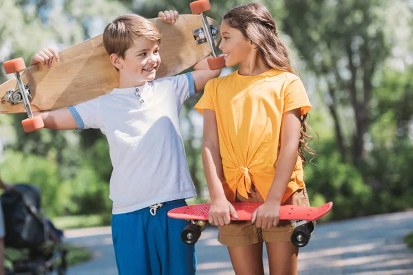 Lindos niños felices sosteniendo patinetas y sonriéndose en el parque - foto de stock