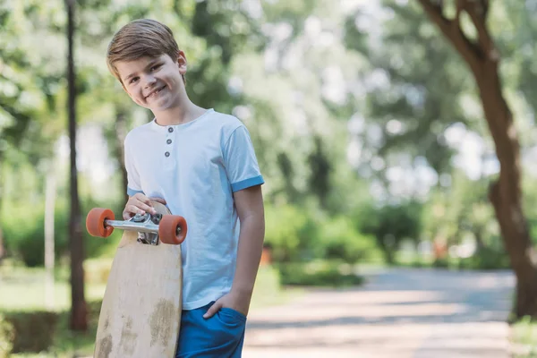 Bambino felice in piedi con skateboard e sorridente alla fotocamera nel parco — Foto stock