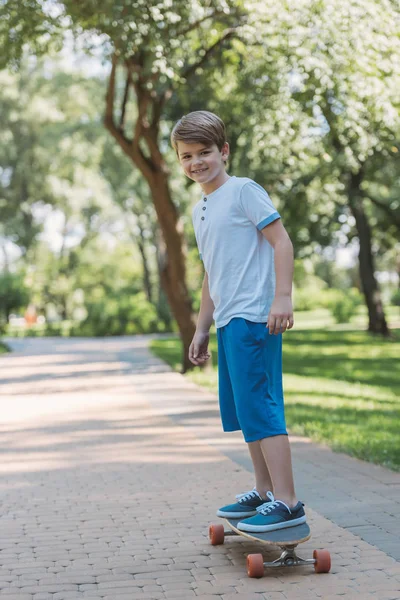Симпатичный счастливый мальчик катается на скейтборде и улыбается фотоаппарату в парке — стоковое фото