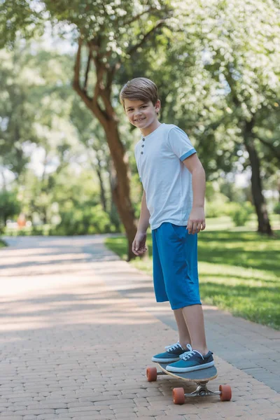 Lindo niño feliz monopatín y sonriendo a la cámara en el parque - foto de stock