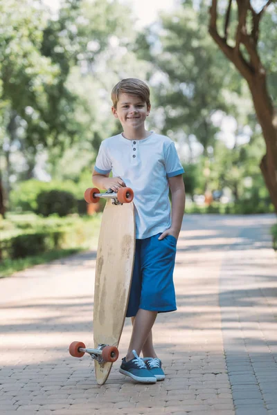 Lindo niño feliz de pie con longboard y sonriendo a la cámara en el parque - foto de stock