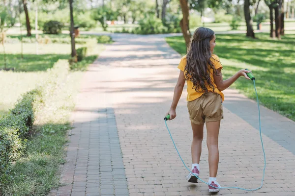 Rear view of cute kid holding jumping rope and walking in park — Stock Photo