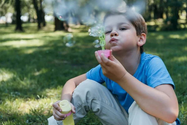 Entzückendes Kind pustet Seifenblasen, während es im Park auf Gras sitzt — Stockfoto