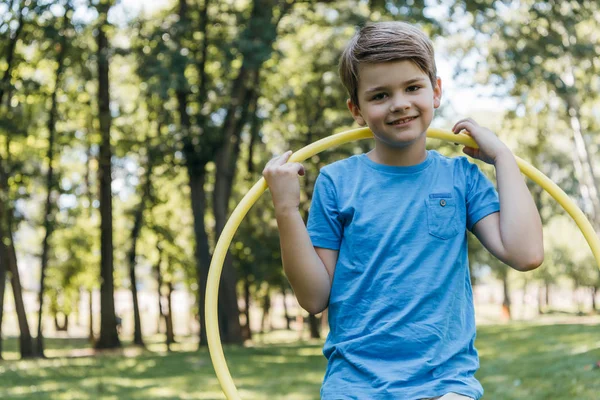Очаровательный счастливый мальчик держит обруч хула и улыбается перед камерой в парке — стоковое фото
