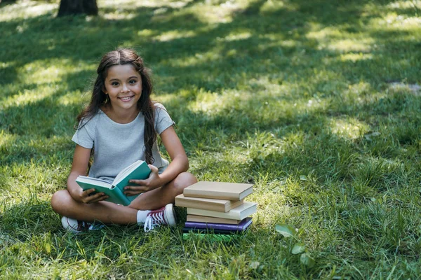 Lindo niño feliz sentado en la hierba con libros y sonriendo a la cámara en el parque - foto de stock