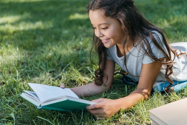 Niedliches lächelndes Kind liegt im Gras und liest Buch — Stockfoto