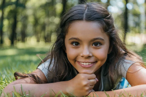 Adorabile bambino felice sdraiato sull'erba e sorridente alla fotocamera nel parco — Foto stock