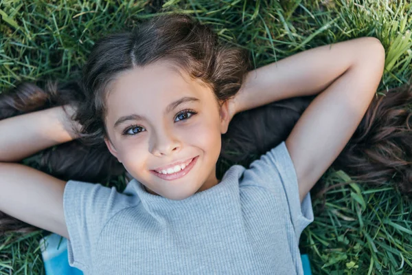 Bellissimo bambino felice sdraiato con le mani dietro la testa sull'erba e sorridente alla macchina fotografica — Foto stock