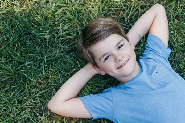 Draufsicht auf einen glücklichen Jungen, der mit den Händen hinter dem Kopf auf dem Gras liegt und in die Kamera lächelt — Stockfoto