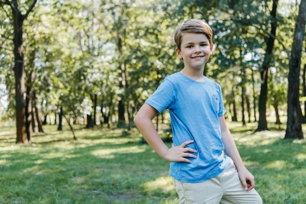 Портрет счастливого мальчика, стоящего с рукой на талии и улыбающегося перед камерой в парке — стоковое фото