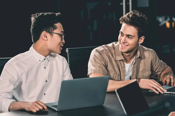 Hombres de negocios multiculturales que trabajan y hablan en la mesa con ordenadores portátiles en la oficina moderna - foto de stock